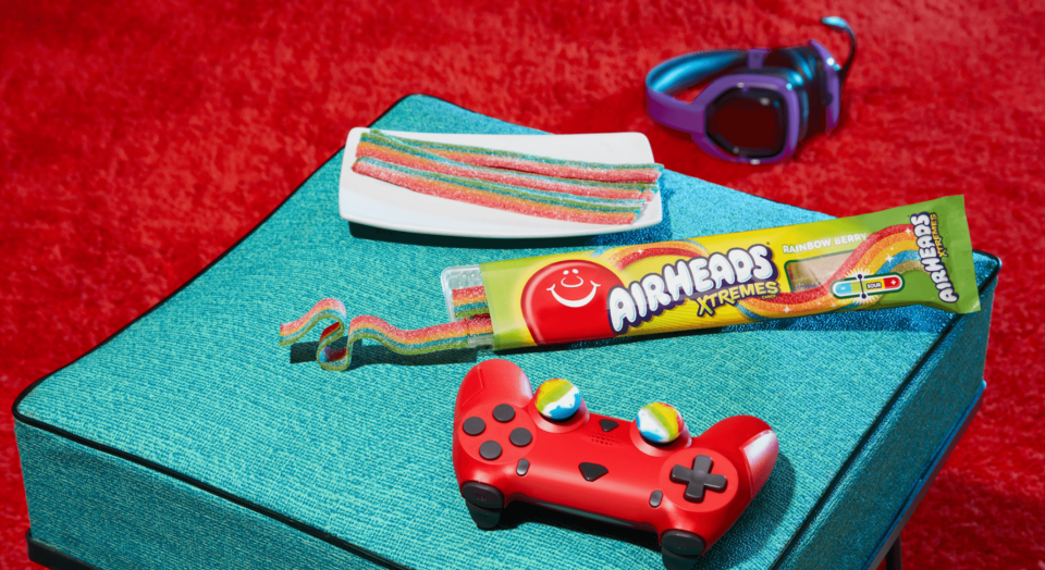 Airheads Xtremes coming out of the package next to a video game controller sitting on a stool with headphones on a red carpet in the background.